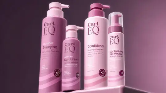 Read the article: Hair Care Products Every Curl Routine Needs Image: CurlEQ shampoo, curl cream, conditioner, and curl defining hair mousse bottles displayed together on a pink studio set