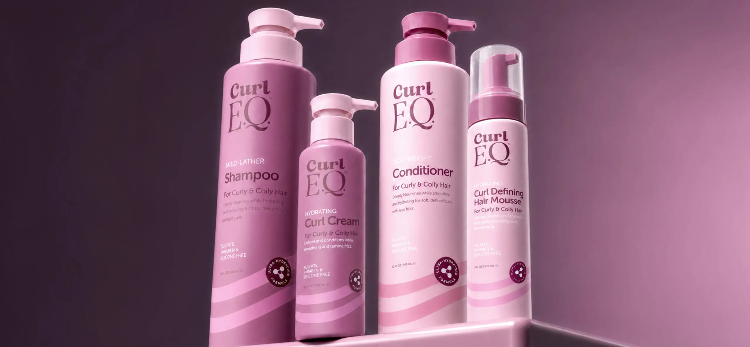 Read the article: Hair Care Products Every Curl Routine Needs Image: CurlEQ shampoo, curl cream, conditioner, and curl defining hair mousse bottles displayed together on a pink studio set